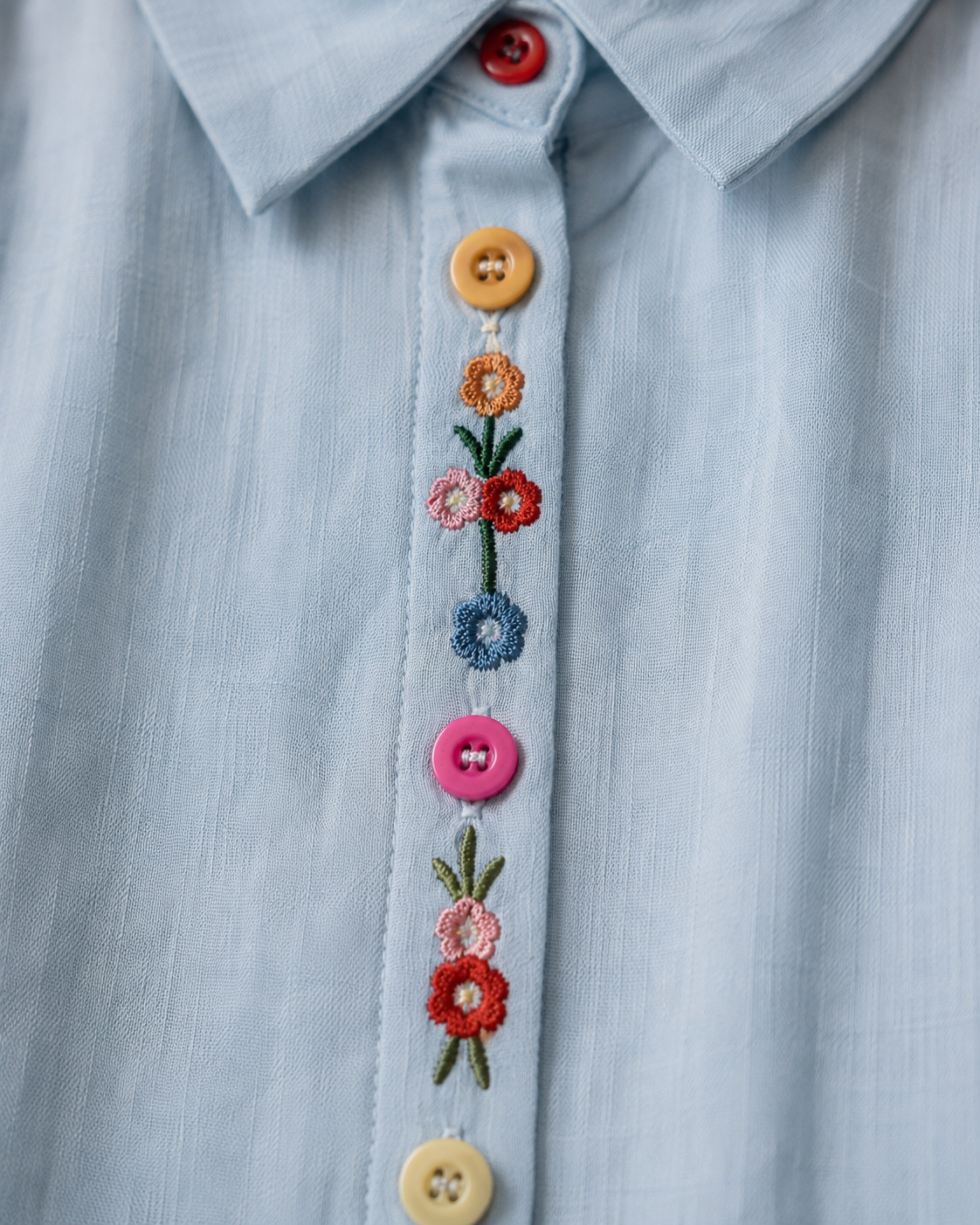 Close-up of wildflower embroidery and decorative buttons on the front placket of the sky blue cotton shirt.
