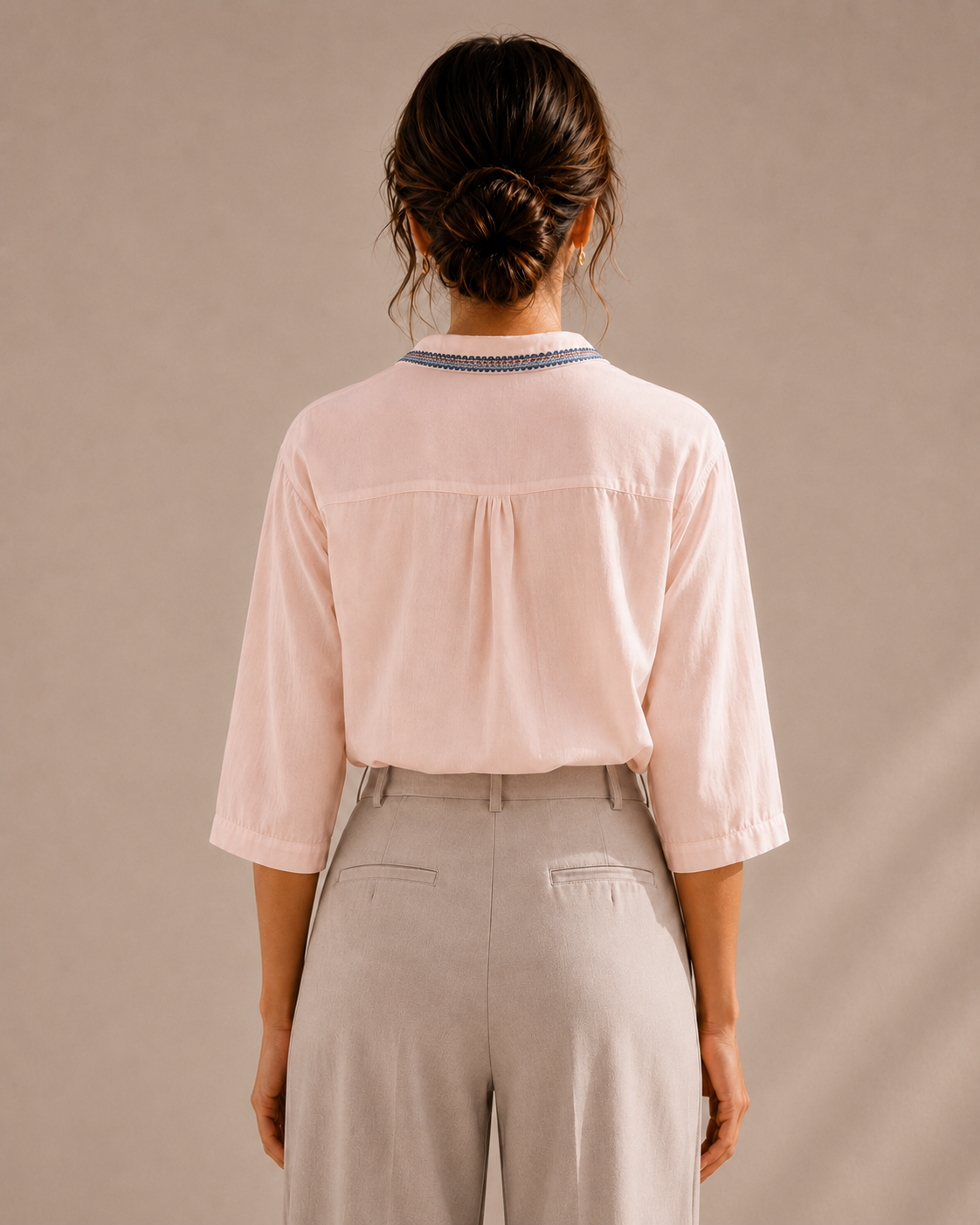 Back view of the soft blush pink medallion embroidered cotton shirt tucked into light trousers, showing the yoke and collar.
