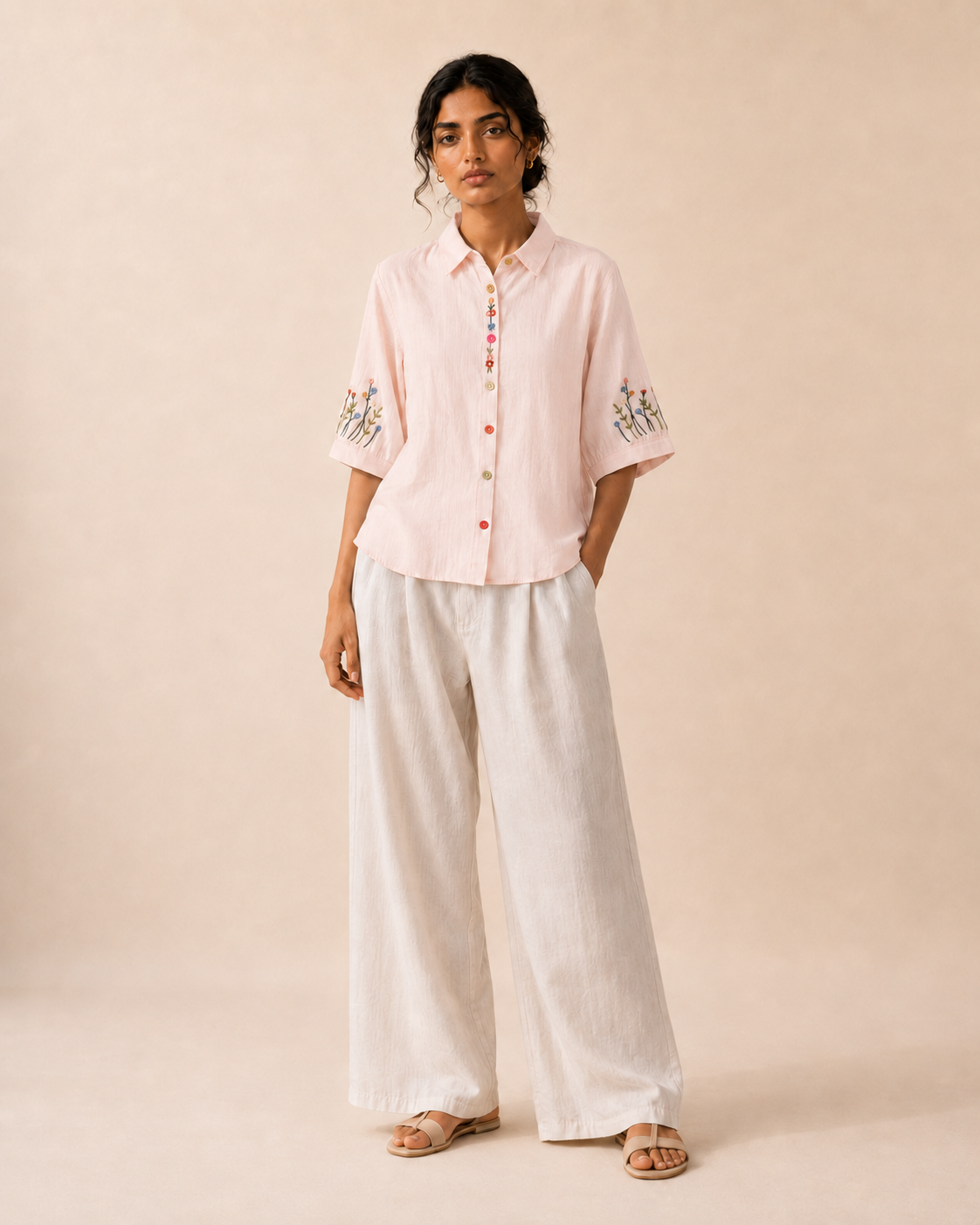 Full-length look of a woman wearing the soft blush pink wildflower embroidered cotton shirt with light trousers.

