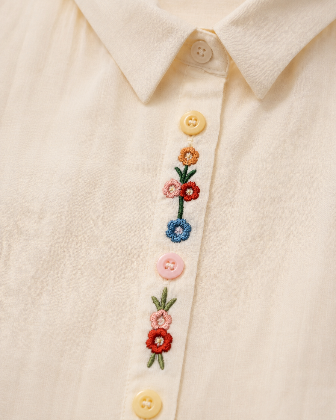 Close-up of wildflower embroidery and decorative buttons on the front placket of the ivory cotton shirt.
